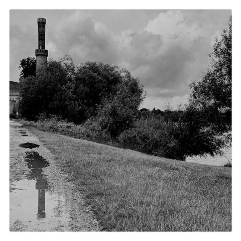 links ein Turm mit Spiegelbild in einer Pfütze, rechts Rasen und Gebüsch, darüber Himmel mit Wolken