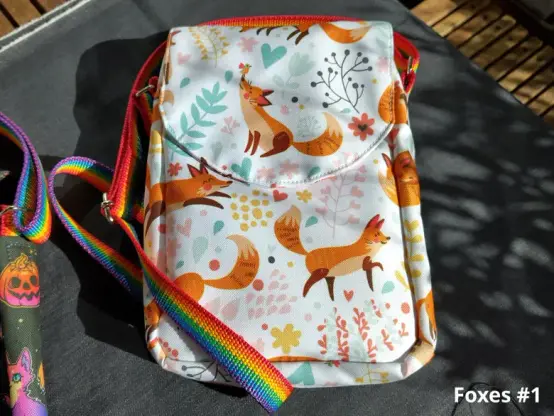 A small shoulder bag made from a white fabric with stylized orange foxes printed on it. The bag has a colorful rainbow strap.