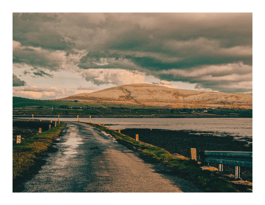 The photo is of a narrow, wet road stretching towards a calm body of water (the Atlantic ocean) and curling to the left of the image. Beyond the water, a low, pale-colored hill is prominent and above it there is a dark, moody, cloudy sky. There are small, evenly spaced yellow arrow signs along the sides of the road.
