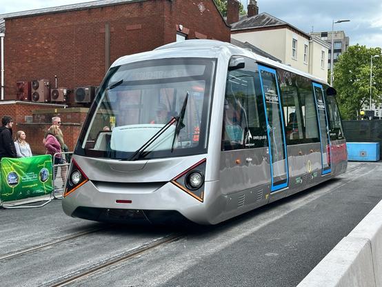 The Coventry Very Light Rail vehicle standing at the top end of the demonstration track in Coventry city centre. It is a small battery tram in silver with light blue doors.