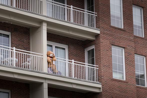 Looking up at a brick building with lots of balconies. One has a large, plush dog hanging on the railing watching the street below.