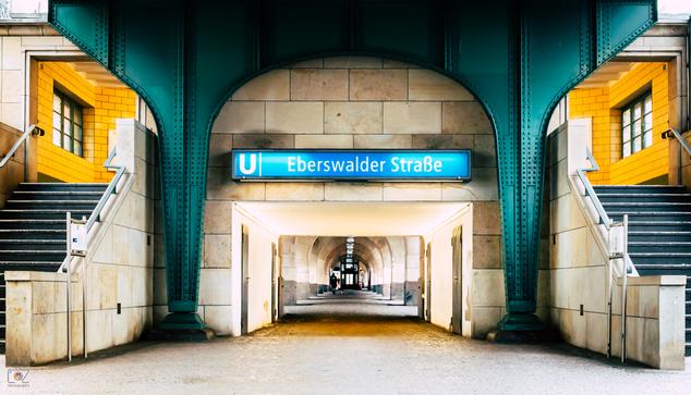 View of the entrance to the "Eberswalder Straße" subway station in Berlin. The architecture is very symmetrical. The rectangular passageway is in the middle and at ground level at camera height. Behind it is a colonnade with a door at the far end. Above the entrance is a blue sign with the white inscription "U Eberswalder Straße". The passageway is framed on both sides by turquoise-green metal struts with rivets, which support the (invisible) rails above. 

Staircases can be seen symmetrically arranged on the left and right at the edge of the photo, the walls of which are tiled in yellow on the upper floor. All other walls and the wall along the stairs are covered with large tiles in beige tones.