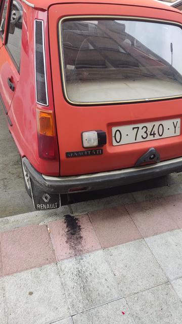 La imagen muestra la parte trasera de un Renault 5 TL rojo estacionado en una acera. El coche tiene una matrícula española que comienza con "0-7340-Y".  Se observa una mancha oscura en el suelo debajo del coche, posiblemente aceite o líquido de escape, y una colilla de cigarrillo cerca. El parachoques y la carrocería del coche muestran señales de desgaste.