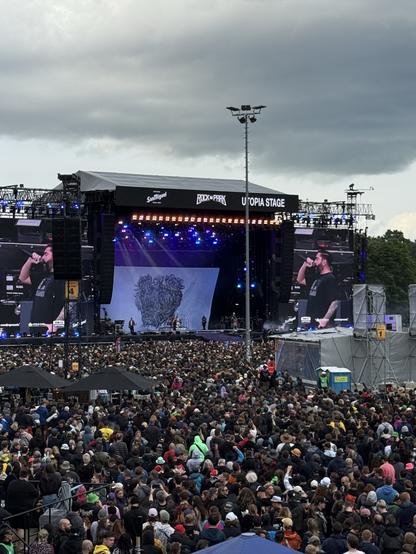A large crowd gathers at the Utopia Stage during a music festival, with a performer visible on stage. The backdrop features an artistic design, and the atmosphere appears energetic with various fans engaging with the performance. The sky is overcast, hinting at