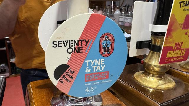 A circular beer tap handle featuring the label for "Tyne & Tay" brewed by Full Circle Brew Co. It includes design elements in blue, red, and black, with descriptions indicating it is a marmalade best bitter, amber in color, with 4.5% ABV