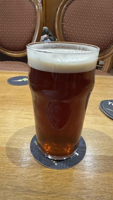 A glass of amber beer with a creamy foam head sits on a wood table. A drink coaster is beneath the glass, and decorative chairs are visible in the background.