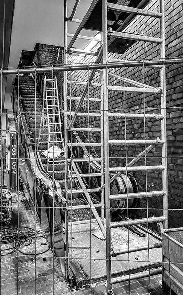 Aufwärts führende Rolltreppe hinter einem Bauabsperrgitter; vor der Treppe steht ein minimales Baugerüst aus zwei Seitenteilen und nur einer Arbeitsebene; auf der Treppe eine Klappleiter, daneben Kabel, Bauschutt,  Pappen – die Fülle von  Einzelheiten erzeugt einen maximal verwirrenden Eindruck.

Escalator leading upwards behind a construction barrier; in front of the staircase is a minimal scaffolding consisting of two side parts and only one working level; on the staircase a folding ladder, next to it cables, building rubble, cardboard - the abundance of details creates a maximally confusing impression.