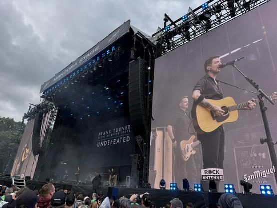 A live music performance on the Mandora Stage featuring Frank Turner and The Sleeping Souls. The artist is singing and playing an acoustic guitar, with band members visible on stage. A large screen displays the performance in front of a crowd, under a cloudy sky