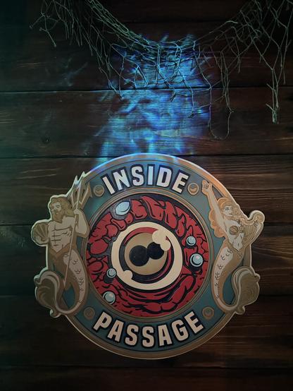 Inside Passage logo: a porthole with "Inside" along the top edge and passage" along the bottom edge. A merman is on the left and a mermaid is on the right. In the center is the eyeball of "Kiki" the  kraken looking out.