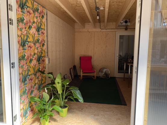 View of the entrance to the workshop office area with flower wall paper and plants, a grey rug and wooden framed chair