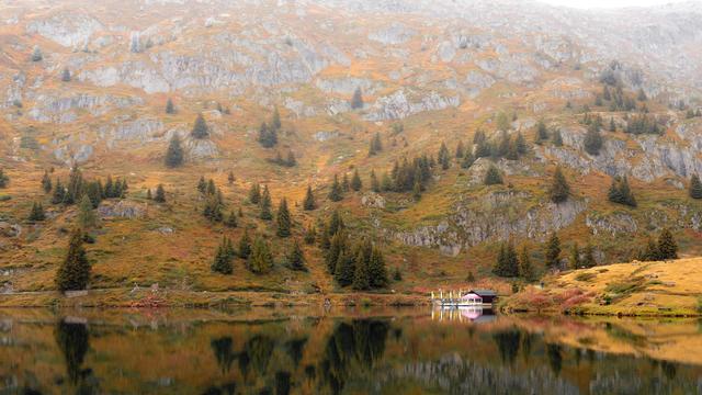 Mountainous landscape with sloping hills covered in mixed vegetation, featuring patches of autumn foliage and scattered evergreen trees. A small cabin is nestled near a calm lake, reflecting the hues of the surrounding scenery. The foreground displays vibrant water reflections, while the mountains rise softly in the misty background.