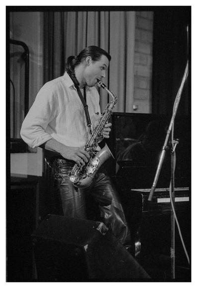 Black and white photograph of a saxophone player. He is seen from the side looking to the right. It looks as if he is communicating with somebody just outside the frame.