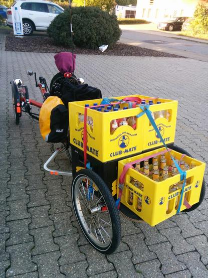 5 leere Matekästen auf einem Fahrradanhänger. 
Der Fahrradanhänger ist an ein Liegedreirad gekoppelt, an dessem Gepäckträger zwei Packtaschen befestigt sind.
Das Foto ist schräg von hinten aufgenommen.