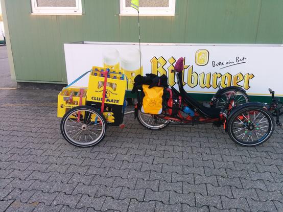 5 leere Matekästen auf einem Fahrradanhänger. Der Fahrradanhänger ist an ein Liegedreirad gekoppelt, an dessem Gepäckträger zwei Packtaschen befestigt sind. 
Das Gespann steht vor einem Getränkemarkt.
Das Foto ist von der Seite aufgenommen.