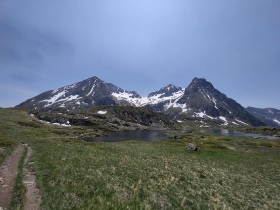 Prairie fleurie, lac et montagne avec 3 sommets encore enneigés, ciel bleu avec juste des lambeaux de nuages.
