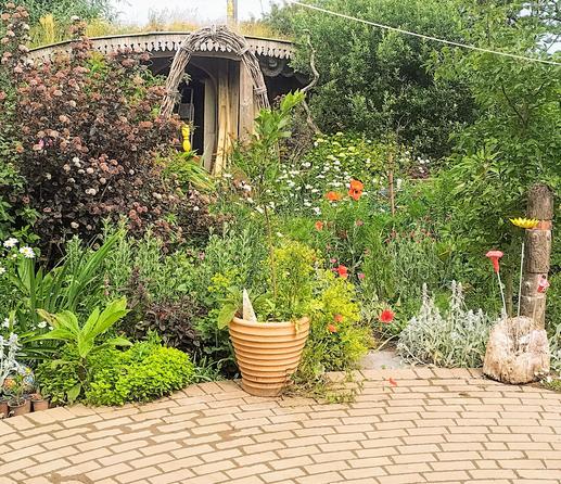 The same area as the previous photo, but taken from a different angle (from rightwards of where the last one was snapped), giving a view more diagonally down the garden. More of one of the sheds is visible, with a flattish green roof and a fascia made of individual leaf-shapes cut out of 4"X1" pieces of timber, and an arch made of branches in front of it.