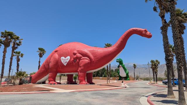 Iconic roadside attraction featuring a massive red brontosaurus statue with "Choose Love" on its heart, and a green T-Rex in the background. The scene is framed by palm trees against a clear blue sky with mountains in the distance.