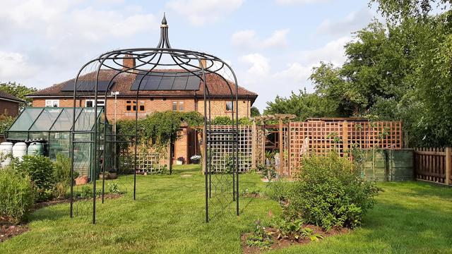Gothic black mild steel gazebo in foreground, although difficult to see it clearly, assembled on lawn and behind it with greenhouse to the left and wooden trellis to the right. Brick house in background with solar panels.