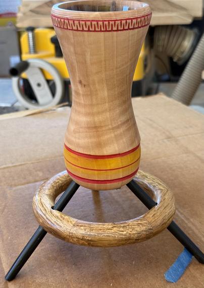 A small vase rests on a tripod of pencil-like pieces of wood which are braced by a ring of wood below the round base of the vase. The vase has yellow and red decorations.