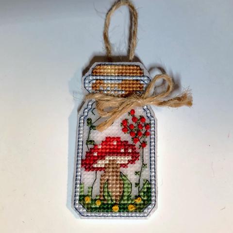 A cross stitch needle point of a small glass jar stoppered with a cork. Inside is a small red mushroom with white spots growing in a grassy spot with small yellow flowers, two vines flank the mushroom, one with small leaves and the other with several small red flowers/berries. There is a twine bow tied around the neck of the jar and a twine loop at the back