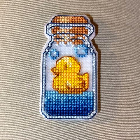 A cross stitch needle point of a small glass jar stoppered with a cork. Inside is a cute, yellow rubber duckie floating  on blue water, there are two bubbles floating by the ducks head