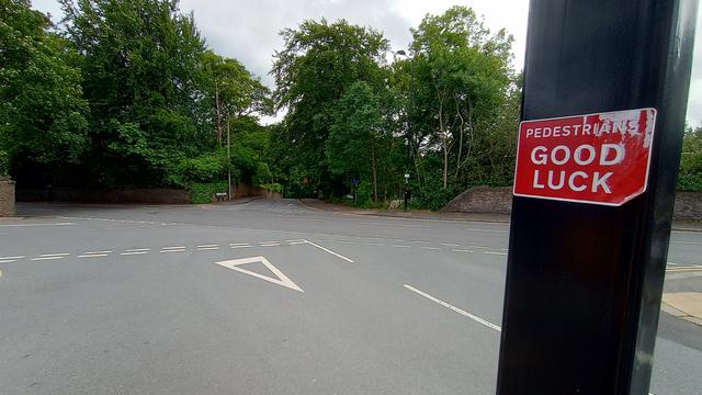 A sticker on a road sign that reads Pedestrians Good Luck, in front of a really badly laid out road junction.