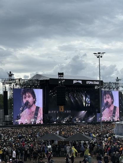 A live concert scene featuring a band performing on the Utopia Stage. There is a large crowd in the foreground, with a big screen displaying the band member singing. The sky is overcast, and some audience members are wearing raincoats. The