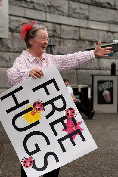This is an image of Marcia on the lower causeway of Victoria's Inner Harbour on June 8, 2010. Exuberant smile in place, Free Hugs poster in hand, and arms open!