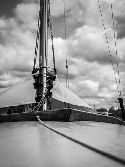 Sailboat deck and wooden mast in black-and-white, with ropes and rigging visible. A taut line extends across the deck. Clouds fill the sky in the background. Valokuvaaja Lappeenranta Kuvaajankulma