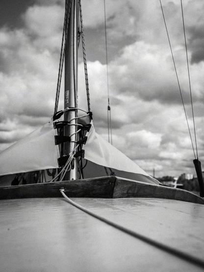 Sailboat deck and wooden mast in black-and-white, with ropes and rigging visible. A taut line extends across the deck. Clouds fill the sky in the background. Valokuvaaja Lappeenranta Kuvaajankulma
