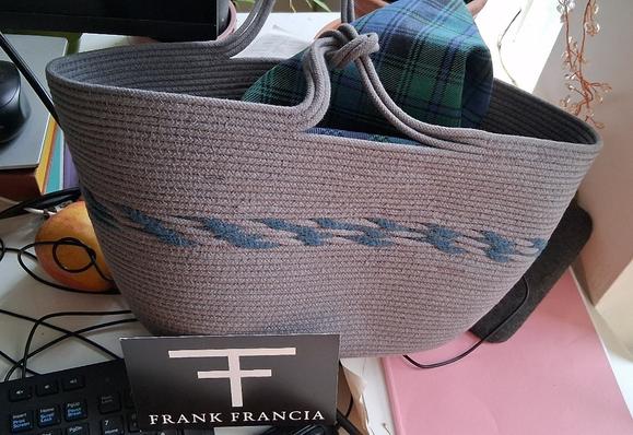 A grey woven bag with knotted handles and blue detailing, with a blue-green tartan pouch inside, resting next to a keyboard on a messy desk, with a card propped up reading FRANK FRANCIA