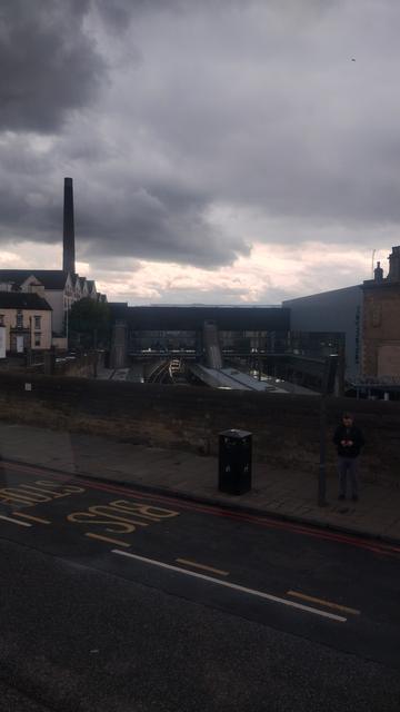 View from the upper deck of a bus, looking over a high, stone wall to the railway station in the cutting below, a mix of bright, white and dark grey clouds above, and an industrial brick distillery chimney on the horizon