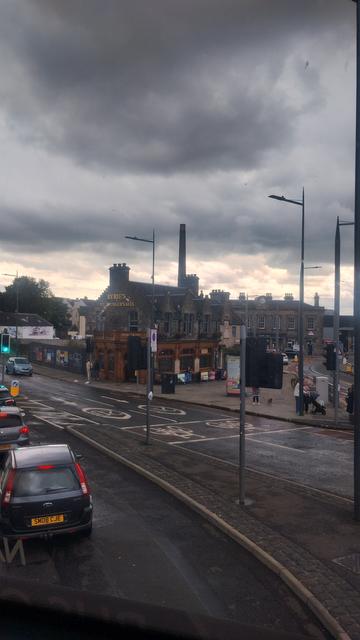View from the upper deck of a bus,showing a Victorian stone built pub, a mix of bright, white and dark grey clouds above, and an industrial brick distillery chimney on the horizon