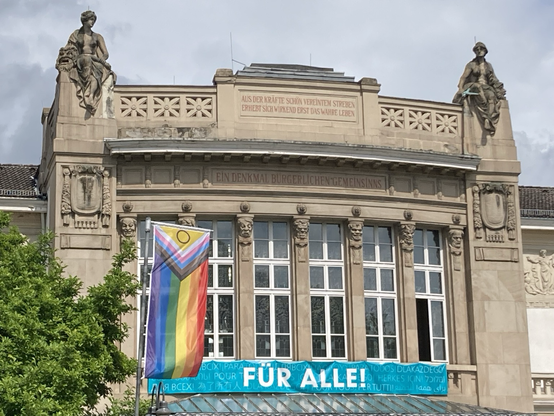 Ansicht des Stadttheaters in Gießen mit einem Banner mit der Aufschrift „Für alle“, an der Fahnenstange daneben weht eine Fahne der LGBTQ - Community mit Transgenderkeil incl der Farben Schwarz und Braun