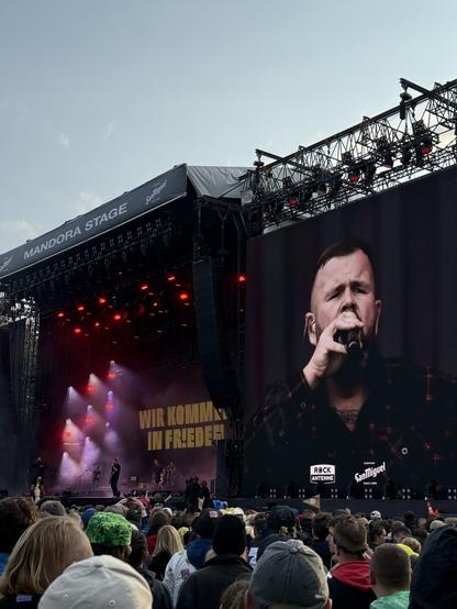 A live music performance at the Mandora Stage with a crowd in front. A singer is prominently featured, performing on stage, accompanied by a band. The background displays the text "WIR KOMMEN IN FRIEDEN." The atmosphere includes stage lights