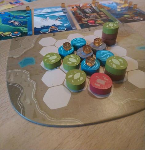 Szene aus dem Spiel "Harmonies". Vorne meine Landschaft die ich mit Holzmarkern gestaltet habe. Man sieht Flüsse, Bäume und Berge. Die besondere Platzierung liefert Punkte. Bestimmte Anordnungen sind zudem gut für Tiere - Zusatzpunkte.