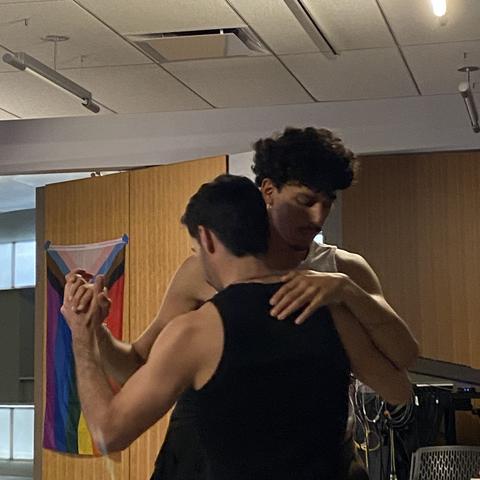Two people with short hair and mustaches wearing tank tops dance chest to chest with a progress pride flag in the background.