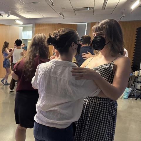 Two masked people, one in a white shirt with hair in bun, the other with loose hair in a checked dress dance in the foreground.