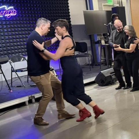 Two short-haired people dance past the stage. One has a black tank top and red boots. Behind them, a woman leads a man in the Americano step