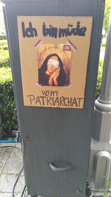 Ein Plakat auf dem steht: Ich bin müde vom Patriarchat. 

Dazu das Bild einer Frau mit langen schwarzen Haaren. Sie trägt eine Augenbinde und hat beide Hände flach zusammen gelegt auf ihrer Wange. Dazu den Kopf leicht geneigt.