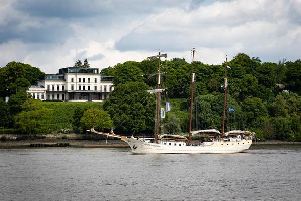 A classic white sailing ship named "Mare Frisium" is cruising along a river, with tall masts and rigging visible against a backdrop of lush green trees. In the background, a large, elegant white mansion stands atop a hill, surrounded by greenery under a partly cloudy sky.