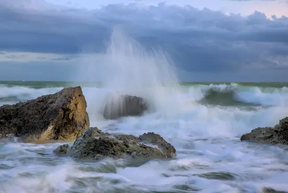 bild im quervormart:
meeresbrandung, am strand liegen mehrere große, grobe felsen, an dem sich das meer bricht. das bild ist mit längerer belichtungszeit geschossen, sodass die gischt weicher ist