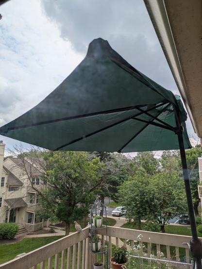 A half-umbrella against a wall on a balcony overlooking a green courtyard with a cloudy summer sky behind. Along the railing, some potted plants and a mourning dove.