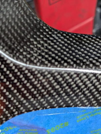Close up shot of a section of carbon fiber during the refinishing process. Visible is a bit of blue painters' tape protecting an edge from over spray.