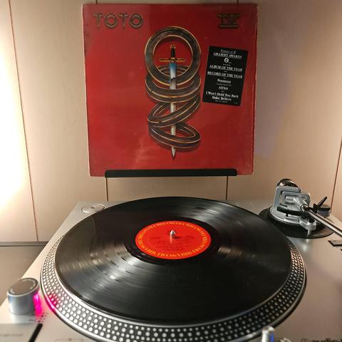 A vinyl record sits on a turntable. Behind the turntable, a vinyl album outer sleeve is displayed. The front cover shows 4 rings around a sword.