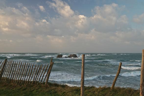 View from atop of a coastal dune down onto a rough sea. In the foreground, there's some short dune grass and a bit of wooden fence in questionable condition. Behind that, the sea extends to the horizon at roughly the vertical centre of the image. There are some large rocks in the water in the centre of the image. There are waves crashing against the rocks and rolling towards the beach, lots of spray can be seen on the water surface throughout. The top half of the image is a cloudy sky with some blue showing. Everything is bathed in warm winter afternoon sunlight.