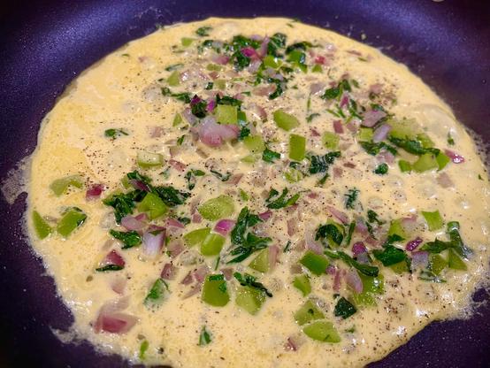 Flat disc frying in a pan that resembles an omelette. There are lots of diced onions, bell peppers, and spinach mixed in.