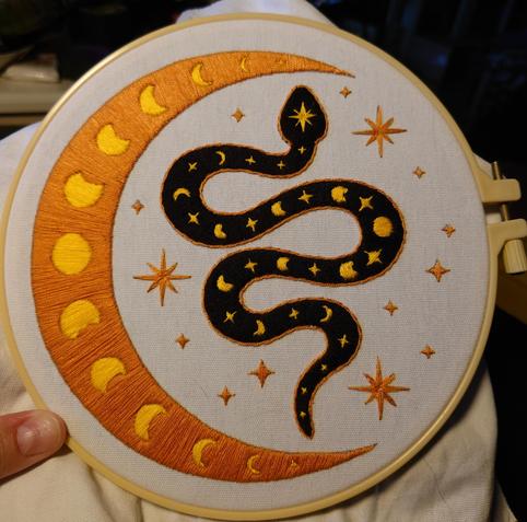 An emboidery in a hoop, with a black snake encircled by a crescent moon. Both snake and moon has a series of moon phases in yellow along them.