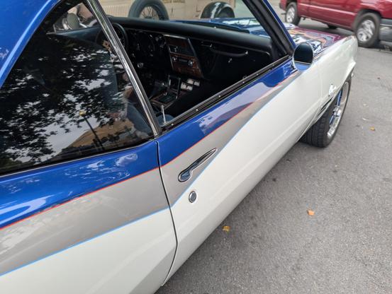 3/4 perspective picture showing the passenger side a first generation Camaro. The side view shows a show quality paint job in a questionable tri-tone with pinstripes. Centered in the shit is a door handle polished to an absolute mirror finish, it has been recessed into the door's outer skin to form a perfectly smooth surface betraying only the shape of the door handle against the contour of the surrounding panel.
