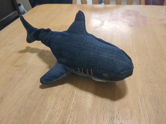 A denim stuffed shark on a wooden table. The body of the shark is dark blue denim, and the tummy is light blue. The eyes and gills are embroidered with gold jeans top-stitching thread, which has also been used to edge-stitch on the fins.